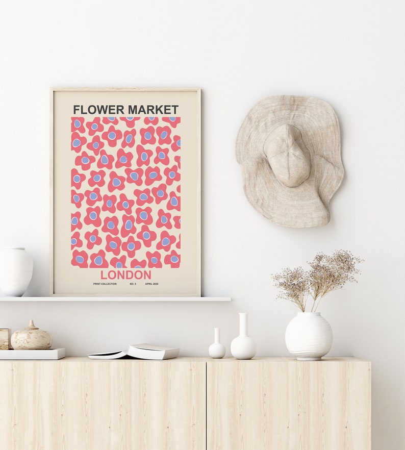 Pu&ograve; includere: Una stampa incorniciata con un motivo floreale rosa e blu e il testo "FLOWER MARKET LONDON" in nero. La stampa &egrave; appesa a una parete bianca sopra uno scaffale in legno con un vaso bianco, un piccolo vaso bianco e una zucca marrone.