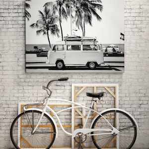 May include: Black and white photograph of a vintage van parked on a beach with palm trees in the background. A white bicycle is in the foreground.