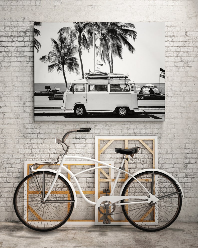 May include: Black and white photograph of a vintage van parked on a beach with palm trees in the background. A white bicycle is in the foreground.