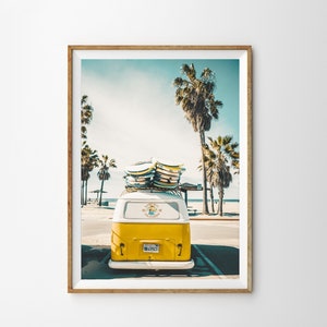 May include: A yellow vintage van with surfboards on the roof parked in front of a beach with palm trees. The van has a white stripe and a sticker that says "Santa Cruz California".