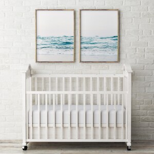 May include: Two framed photographs of ocean waves hanging above a white crib with a white mattress.