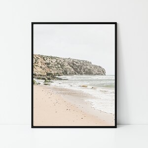 May include: Framed print of a coastal landscape. The image features a sandy beach with gentle waves, a rocky cliff, and a cloudy sky. The color palette is muted, with beige, gray, and green tones. The artwork is displayed in a black frame.