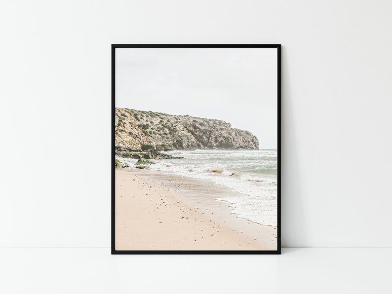 May include: Framed print of a coastal landscape. The image features a sandy beach with gentle waves, a rocky cliff, and a cloudy sky. The color palette is muted, with beige, gray, and green tones. The artwork is displayed in a black frame.
