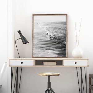 May include: A black and white photograph of surfers riding waves. The photo is framed in a brown wood frame and is hanging on a white wall above a wooden desk with a white drawer and a black metal stool.