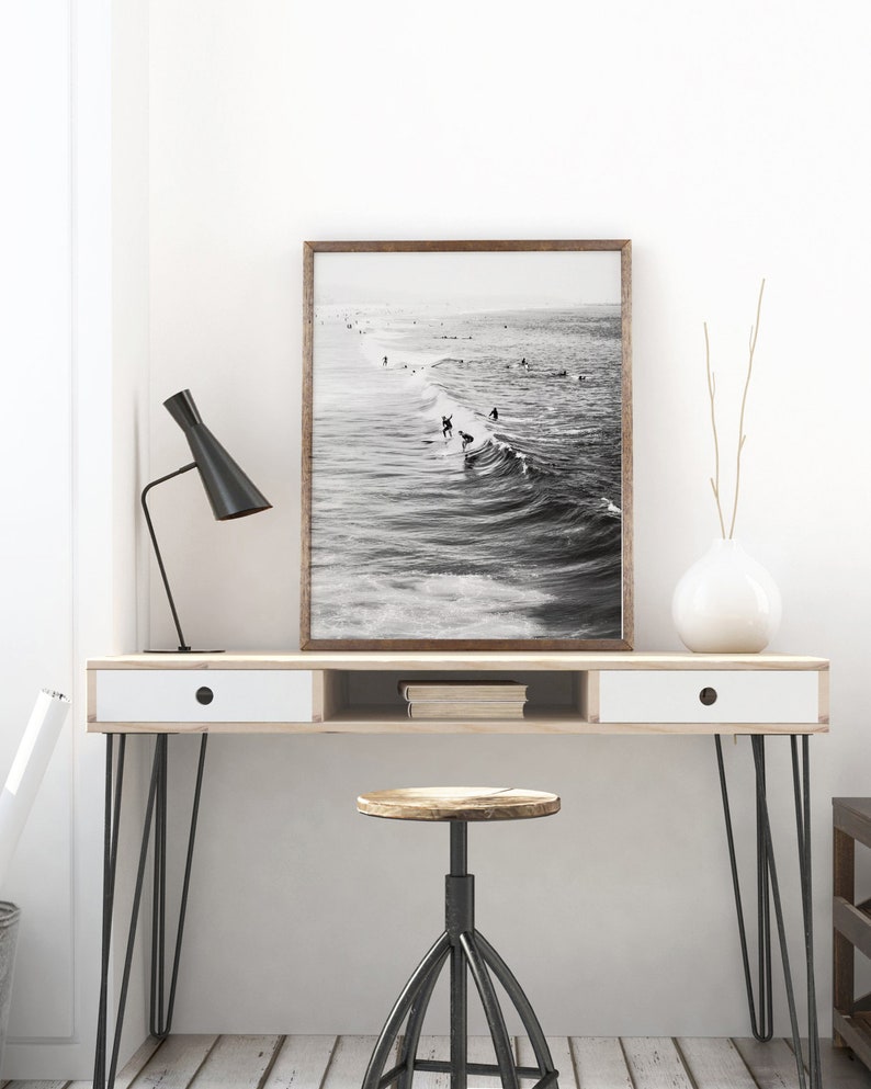 May include: A black and white photograph of surfers riding waves. The photo is framed in a brown wood frame and is hanging on a white wall above a wooden desk with a white drawer and a black metal stool.