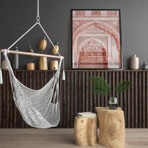 May include: A white macrame hammock chair hangs from the ceiling in front of a dark wood slat wall. A framed print of an intricate archway in red tones hangs above a wooden shelf with decorative objects. Two wooden stools with natural bark finishes sit in front of the wall.