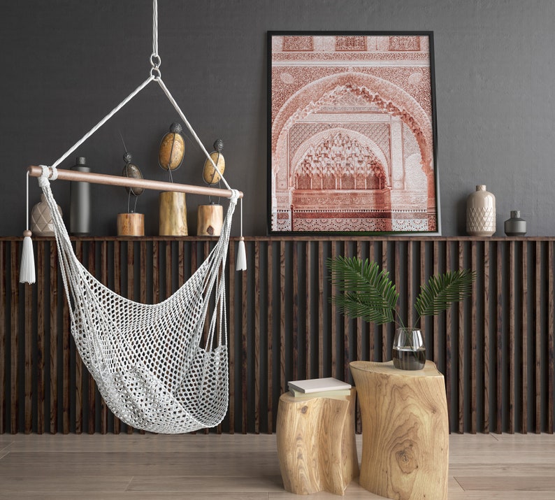 May include: A white macrame hammock chair hangs from the ceiling in front of a dark wood slat wall. A framed print of an intricate archway in red tones hangs above a wooden shelf with decorative objects. Two wooden stools with natural bark finishes sit in front of the wall.
