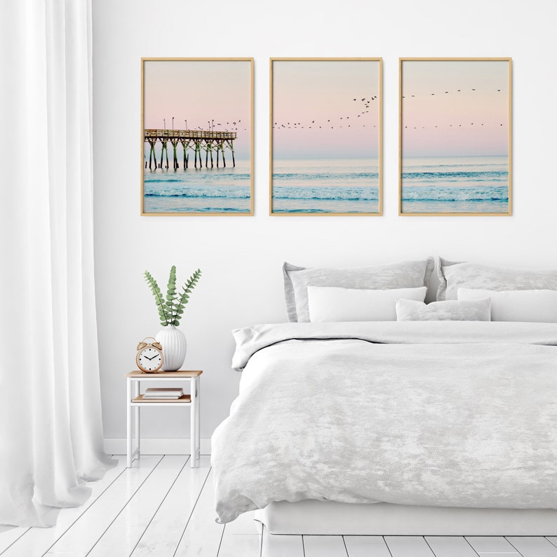 May include: Three framed photographs of a beach scene with a pier and birds flying overhead. The photos are hung on a white wall above a white bed with a white duvet cover and white pillows.