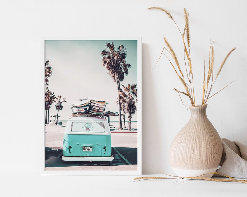 May include: A vintage teal blue van with surfboards on the roof parked on a beach with palm trees in the background.