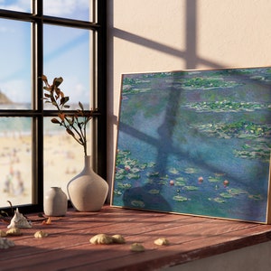 May include: A framed painting of water lilies in shades of blue and green, with a few pink flowers visible. The painting is hanging on a wall next to a window with a view of a beach.