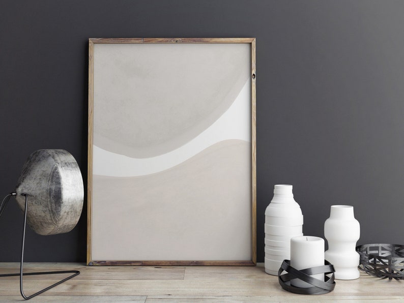 May include: A framed abstract print with beige and white watercolour-like shapes hangs on a dark grey wall.  A silver lamp, two white ceramic vases, and a black candle holder with a white candle are on a wooden table in front of the print.