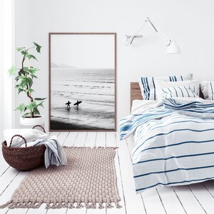 May include: A white bedroom with a bed with blue and white striped bedding. There is a black and white photo of two surfers on the wall above the bed. A potted plant is in the corner of the room. A rug is on the floor in front of the bed.