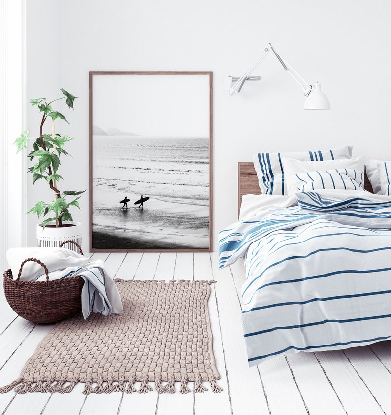May include: A white bedroom with a bed with blue and white striped bedding. There is a black and white photo of two surfers on the wall above the bed. A potted plant is in the corner of the room. A rug is on the floor in front of the bed.