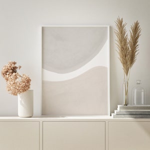 May include: A white framed abstract art print with a beige and grey colour scheme. The print is hanging on a white wall above a white cabinet with two doors. There are dried pampas grasses in a clear glass vase and a clear glass cloche on the cabinet.