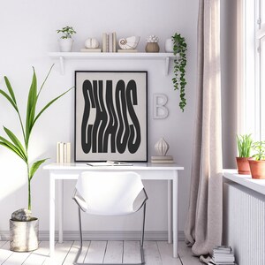 May include: A black and white poster with the word "CHAOS" in a stylized font. The poster is framed in black and hanging on a white wall above a white desk with a white chair. There are plants and decorative objects on a white shelf above the poster.