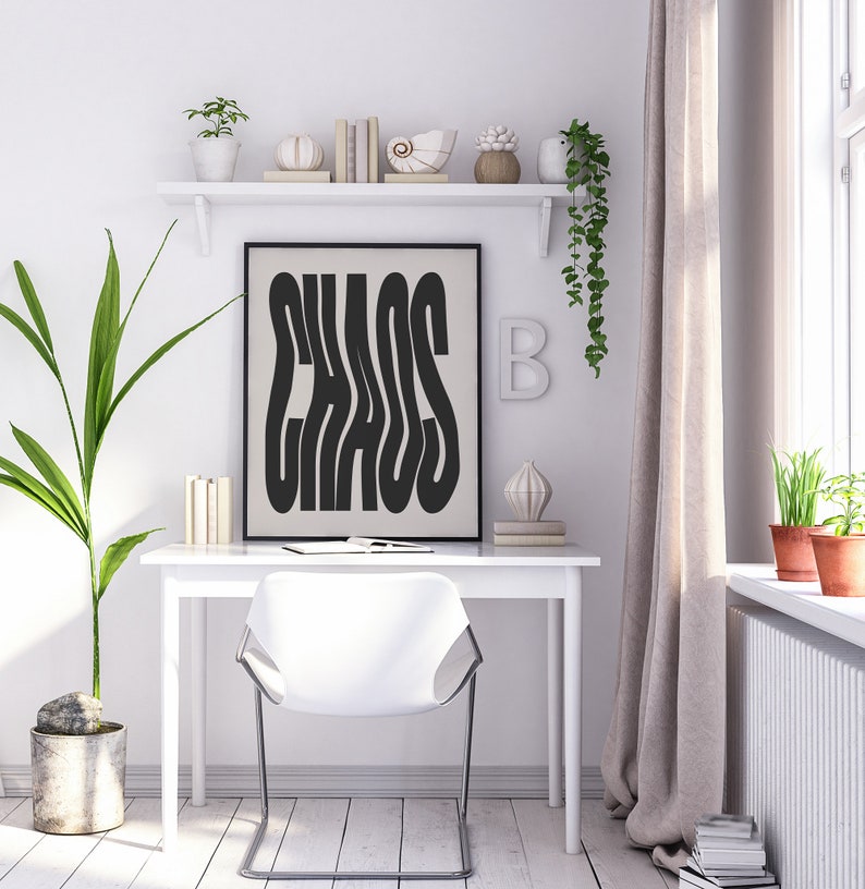 May include: A black and white poster with the word "CHAOS" in a stylized font. The poster is framed in black and hanging on a white wall above a white desk with a white chair. There are plants and decorative objects on a white shelf above the poster.