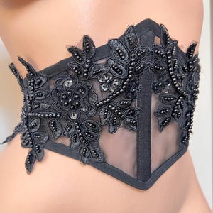 May include: Black lace face mask with floral and beaded embellishments. The mask features a sheer mesh center and is adorned with intricate leaf and flower designs. The edges are trimmed with a dark fabric.