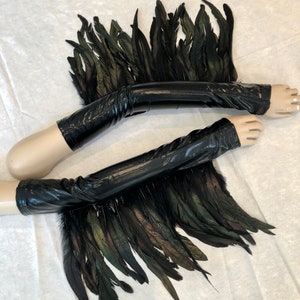May include: A pair of black leather arm warmers with black and green feathers attached. The arm warmers are on a mannequin arm.