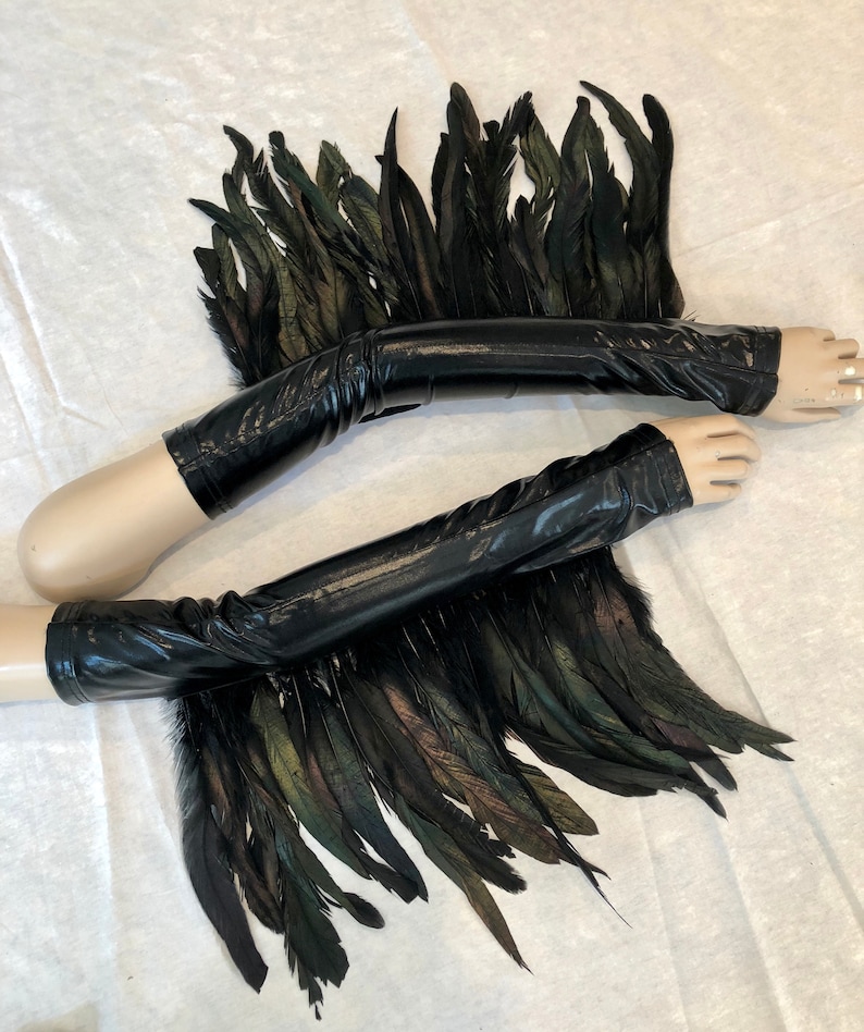 May include: A pair of black leather arm warmers with black and green feathers attached. The arm warmers are on a mannequin arm.