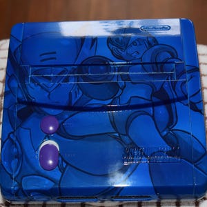 Può includere: Una console Nintendo Super Famicom blu con un design personalizzato con personaggi della serie di videogiochi Mega Man X. La console ha due pulsanti viola e un logo Nintendo.