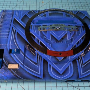 May include: Blue Sega Genesis console with an art deco design. The console has a black circular accent and the text "HIGH DEFINITION GRAPHICS" and "SEGA GENESIS". The power button is red. A unique item for retro gaming fans.