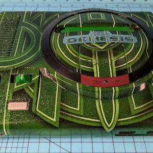 May include: A custom-painted Sega Genesis console in green and white. The console has a geometric design and the word "GENESIS" in silver. The power button is red. A black circular disc is on top.