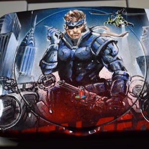 Può includere: Console PlayStation dipinta su misura con un'illustrazione dettagliata di un personaggio in abito blu e nero, ambientata sullo sfondo di un paesaggio urbano. Il design include un elicottero e il logo PlayStation.