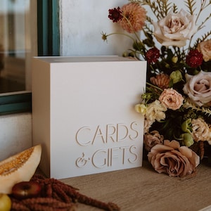 May include: A white, square card and gift box with the words "CARDS & GIFTS" embossed on the front. The box is on a wooden surface, with a floral arrangement of roses and other flowers in the background. A slice of melon and other fruit are in the foreground.