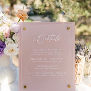 May include: A pink acrylic cocktail menu with gold accents. The menu lists cocktail names and ingredients in white script. The menu is displayed on a white surface with flowers and greenery in the background.
