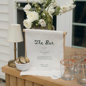May include: A bar setup with a white floral arrangement, a small lamp, and a sign that reads "The Bar." The sign lists cocktail options. Clear glasses with sugared rims are arranged on the wooden surface, along with a bowl of dried citrus.