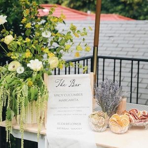 May include: A bar display featuring a sign with "The Bar" written on it, listing cocktails like Spicy Cucumber Margarita and Elderflower Herbal Smash. Dried citrus fruits and lavender are arranged nearby. A floral arrangement of yellow and white flowers is on the left.
