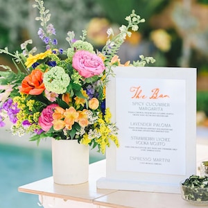 May include: A white sign with the text "The Bar" in orange script. The sign lists a variety of cocktails, including Spicy Cucumber Margarita, Lavender Paloma, Strawberry Lychee Vodka Mojito, and Espresso Martini. A bouquet of colourful flowers in a white vase sits on a table in front of the sign.