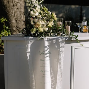 May include: A white linen table runner with lace trim draped over a white bar. The runner features green printed text reading The Bar, Classic Martini, vodka, dry vermouth, bitters, olive, Old Fashioned, whiskey, simple syrup, bitters, BEER | WINE | COCKTAILS.