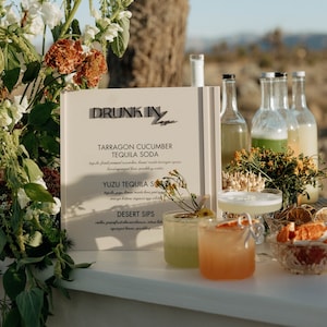 May include: A white sign with the text "DRUNK IN LOVE" and a menu of cocktails including "TARRAGON CUCUMBER TEQUILA SODA" and "YUZU TEQUILA SODA". The sign is on a table with three glasses of cocktails and bottles of liquor.