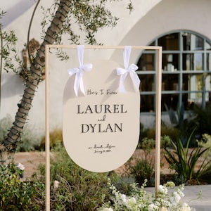 May include: A wooden frame with a white sign that says "Here's To Forever Laurel and Dylan" with the date "June 23, 2023" and the location "San Diego, California". The sign is hanging from white ribbons and is decorated with white flowers.