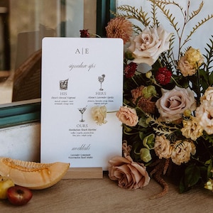 May include: A white sign with the text "AE signature sips" and a list of cocktails with their ingredients. The sign is propped up on a wooden surface next to a bouquet of pink and yellow flowers.