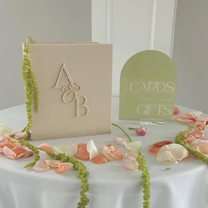 May include: A beige wedding card box with the initials "A & B" and a green sign that says "Cards and Gifts" on a white tablecloth. The table is decorated with flower petals and greenery.