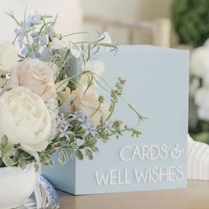 May include: A light blue box with the words "CARDS & WELL WISHES" in white lettering. The box is decorated with a floral arrangement of white, light pink, and blue flowers. The flowers are in a white vase.