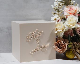 May include: A beige square box featuring the names Kate & Andrew in raised, cursive lettering on the front. The box is positioned next to a decorative arrangement of artificial cream, pink, and mauve roses with green leaves and a small yellow pear.