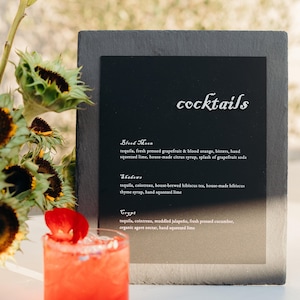 May include: A black chalkboard menu with the word "cocktails" written in white. The menu lists three cocktails: Blood Moon, Shadows, and Trypt. Each cocktail is described with a list of ingredients. A glass of red liquid with a red flower garnish sits on a white surface in front of the menu.