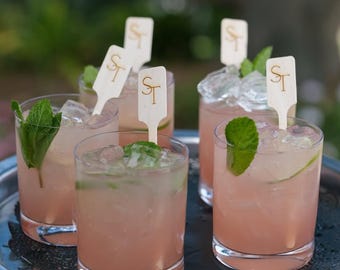 May include: Five clear glass tumblers filled with a light pink cocktail, ice cubes, lime slices, and fresh mint sprigs. Each glass contains a wooden drink stirrer featuring the gold-colored letters ST printed on the top rectangular tab.