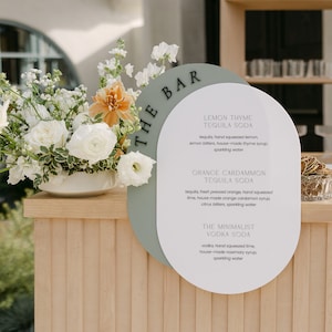 May include: A drinks menu with three cocktail options: Lemon Thyme Tequila Soda, Orange Cardamom Tequila Soda, and The Minimalist Vodka Soda. The menu is white with a sage green accent, and is next to a floral arrangement.