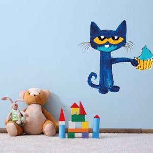 May include: A blue cat with yellow eyes holding a blue and yellow cupcake on a light blue wall. There are stuffed animals and a wooden block tower in the foreground.