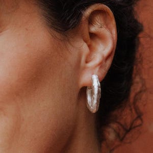 Op de afbeelding: Een vrouw draagt zilveren oorringen. De oorbellen hebben een getextureerd oppervlak en zijn van gemiddelde grootte. De vrouw lacht, met rode lippenstift en bruine ogen. Haar haar is donker en krullend.