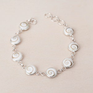 Puede incluir: Un brazalete de plata con abalorios de concha blanca con un patrón en espiral. El brazalete tiene una cadena delicada y es una hermosa pieza de joyería.