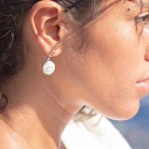 Może przedstawiać: Kobieta o ciemnobrązowych włosach w białej bluzeczce i kolczykach w kształcie muszli.