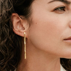 Op de afbeelding: Een vrouw draagt een lange, gouden, getextureerde staaf oorring. De oorring is ongeveer 5 cm lang.