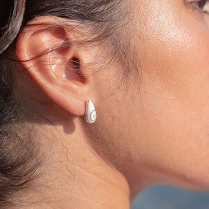 Può includere: Primo piano del viso di una donna, che mostra il suo profilo. Ha i capelli castano scuro e indossa un piccolo orecchino bianco a forma di lacrima.