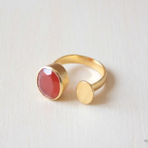 May include: A gold ring with an open design. It features a large, oval-shaped, red gemstone on one end and a small, round gold accent on the other. The ring is set against a light background.