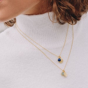 May include: A woman wearing a white turtleneck sweater with a gold chain necklace with two pendants. The pendants are a small round blue stone and a larger round green stone.